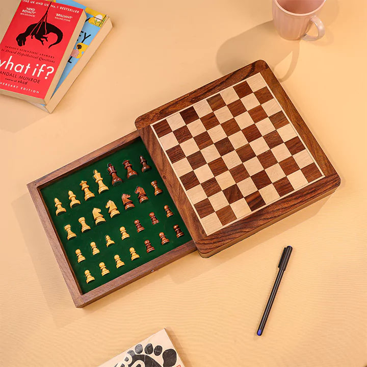 Wooden chessboard with pieces on a beige surface, next to books and a pen.