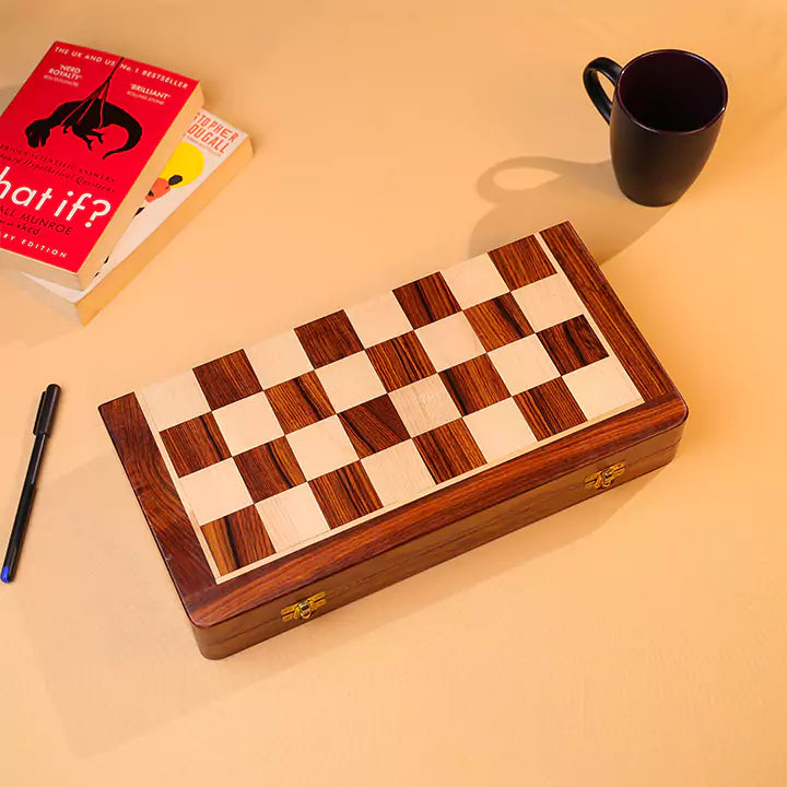 Wooden chessboard with checkered pattern on a beige surface, next to a book and a mug.